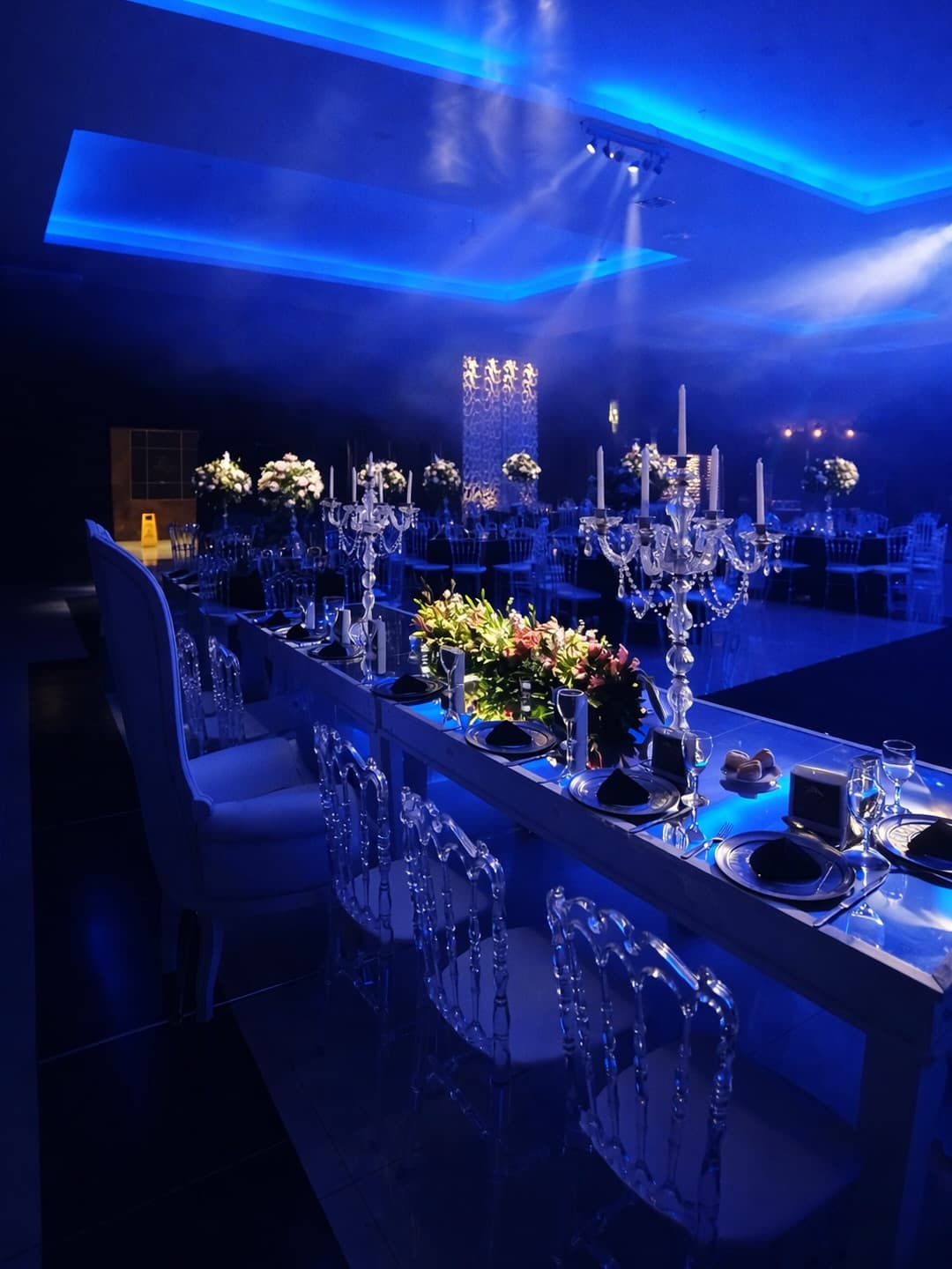 El salón puesto y vacío antes de la fiesta, con la mesa principal de candelabros de cristal y la pista iluminada de azul al fondo.