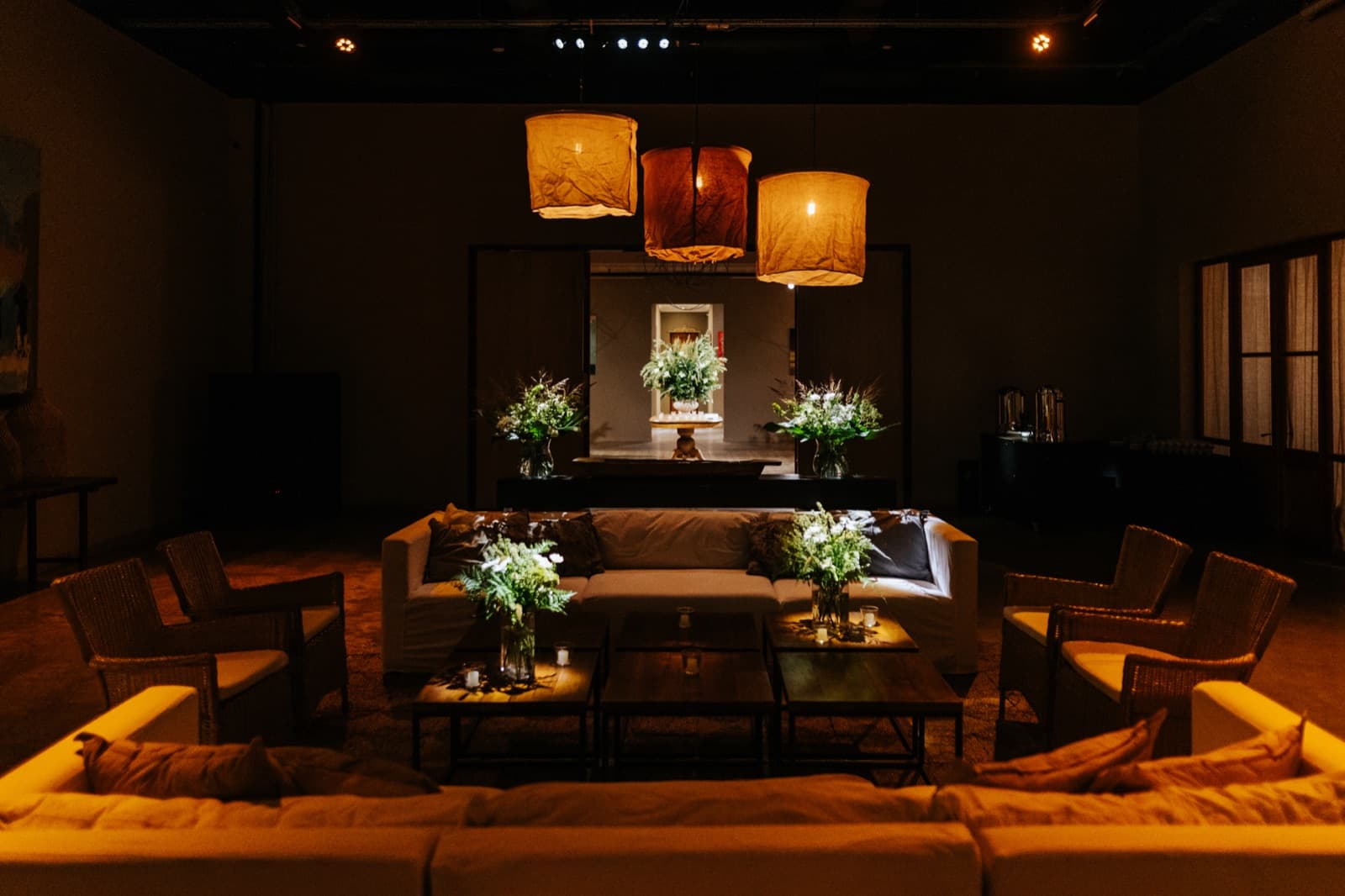 El salón armado como living, con sillones claros alrededor de mesas bajas con velas, tres lámparas grandes de tela colgadas y arreglos de flores blancas al fondo.