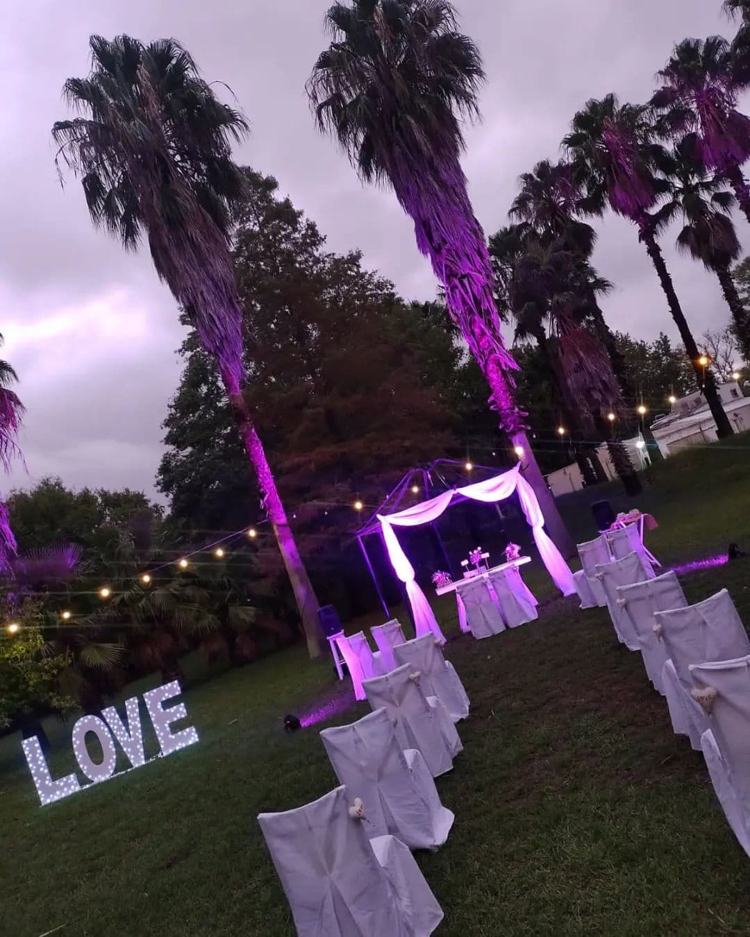 La ceremonia armada en el parque al atardecer: dos hileras de sillas vestidas de blanco frente a un arco con cortinas, las palmeras iluminadas de violeta y unas letras LOVE encendidas sobre el césped.