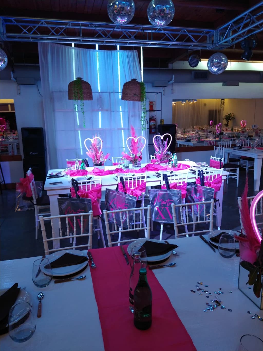 El salón montado para una fiesta de quince, con las mesas largas vestidas de blanco y fucsia, corazones de neón de centro y bochas espejadas colgando de la estructura del techo.