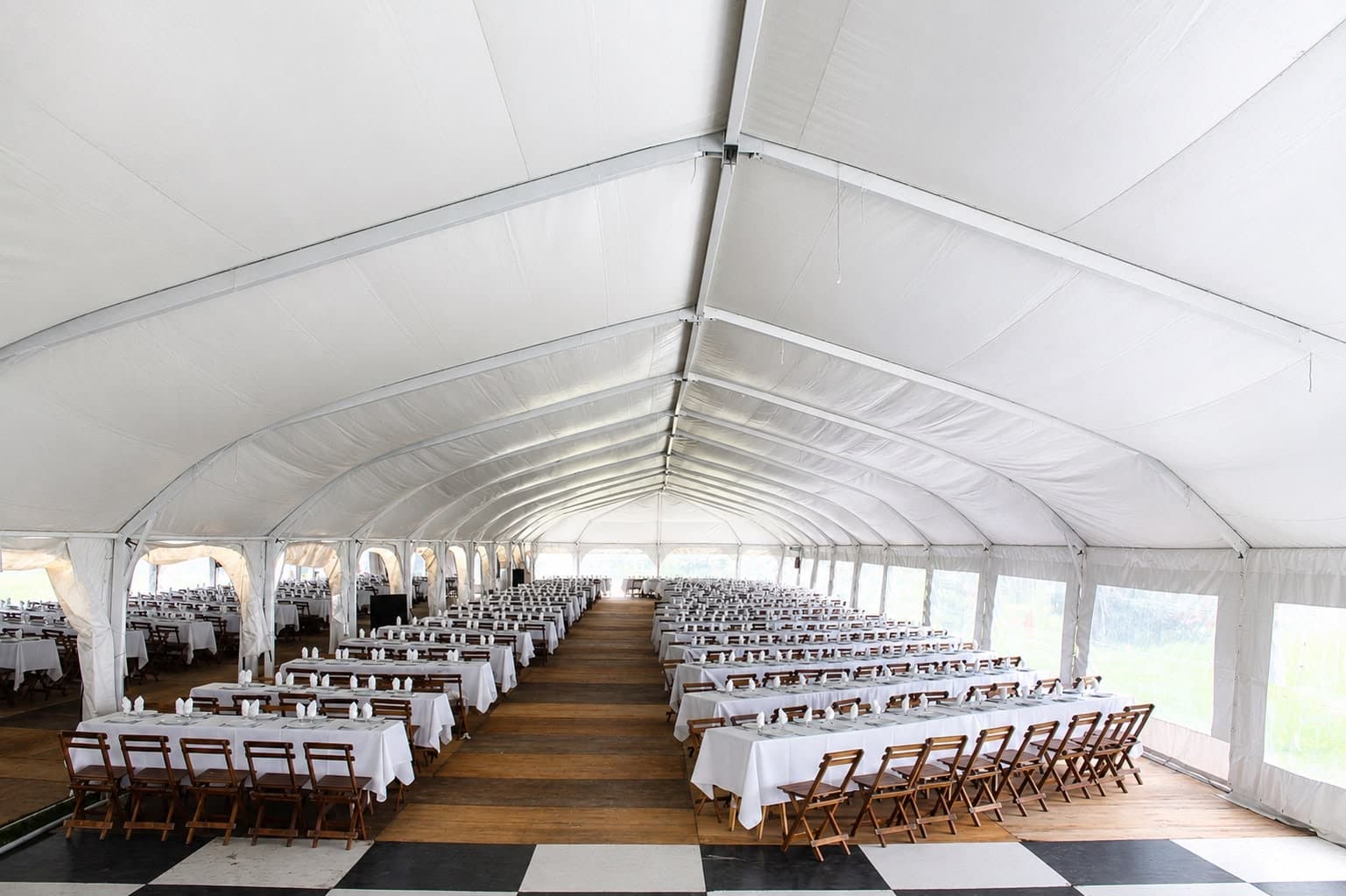 La carpa blanca de estructura arqueada armada para un banquete, con hileras de mesas largas de mantel blanco y servilletas paradas, sillas de madera plegables, una pasarela de tablones en el medio y el piso de damero blanco y negro a los costados.