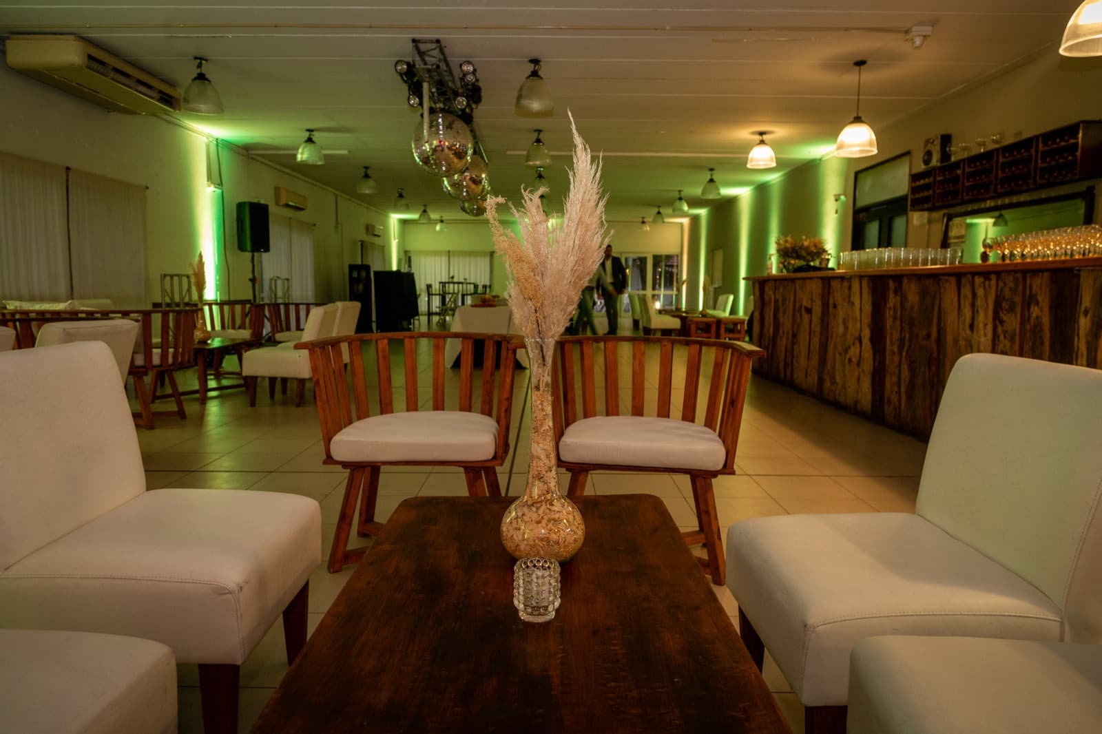 El salón puesto en modo lounge, con sillones blancos y mesas ratonas de madera, una barra larga con frente de troncos y las copas alineadas arriba, las bolas de espejos colgando del techo y las paredes bañadas de luz verde.
