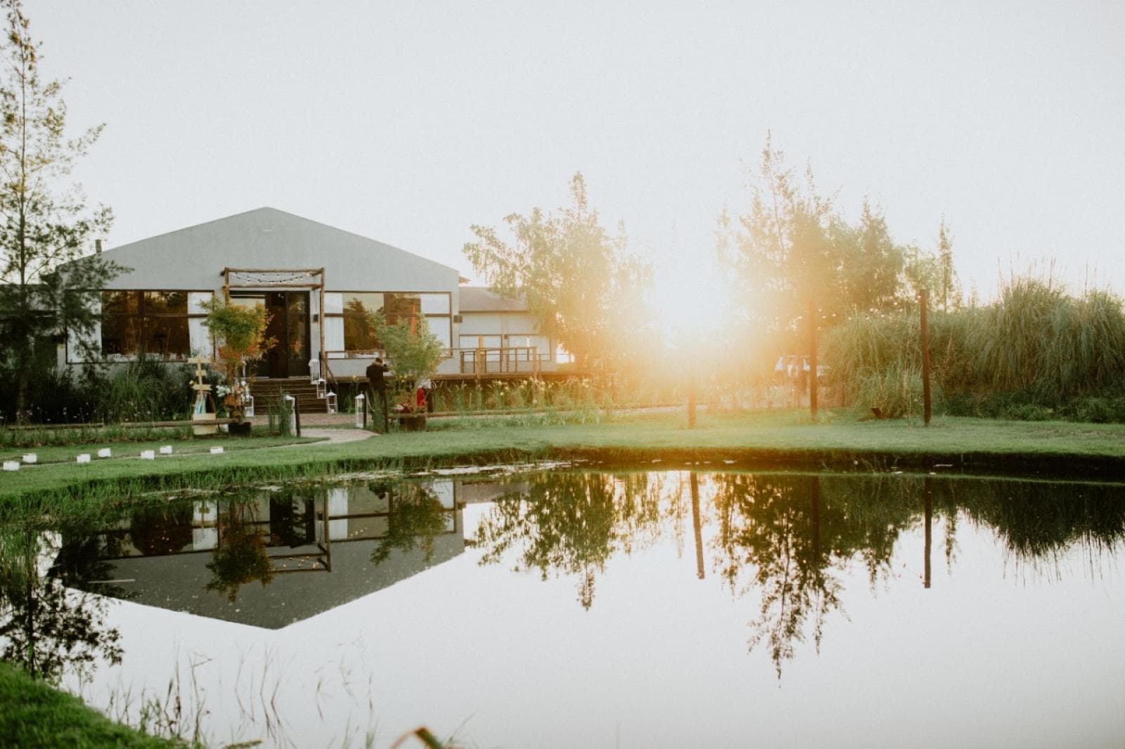 La casa de eventos reflejada en la laguna, con el sol bajo entre los árboles y las plumas de pampa en la orilla.