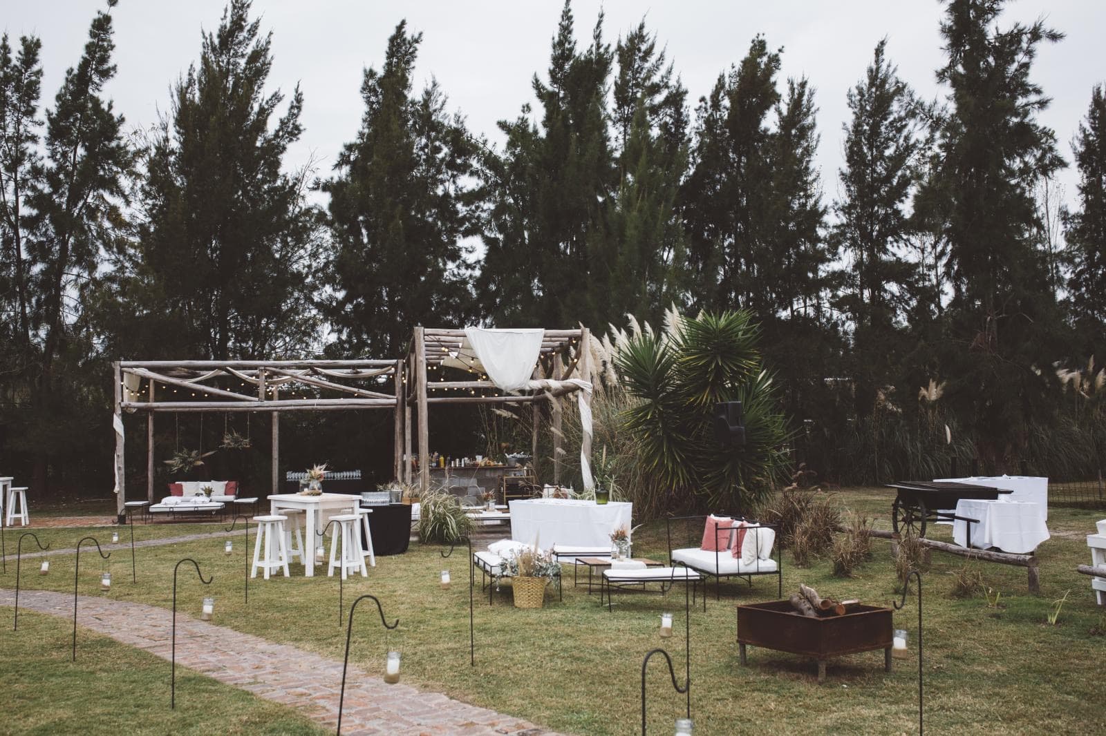 La pérgola de madera con livings blancos, mesas altas y braseros sobre el césped, entre los pinos.