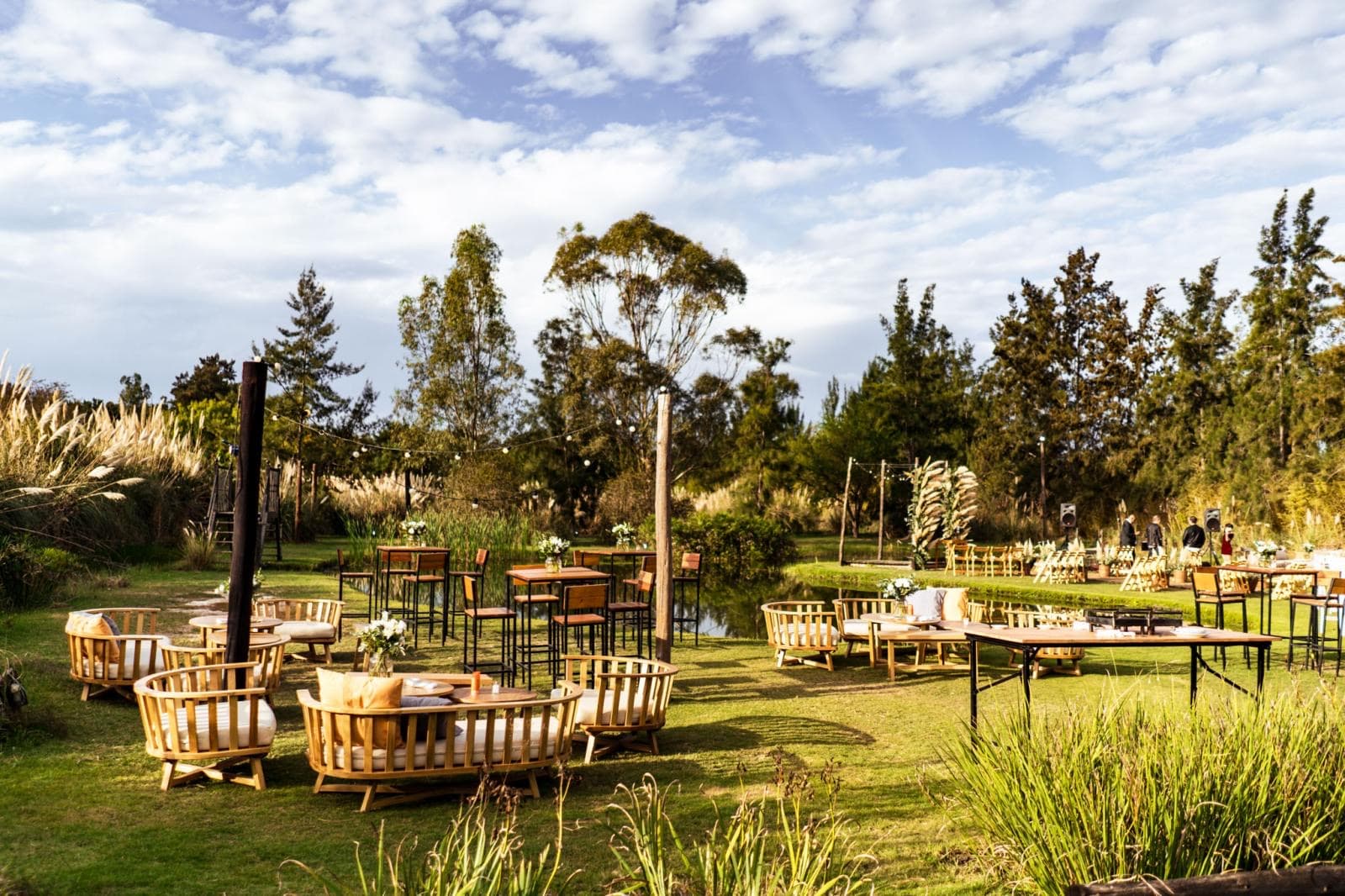 El parque de día con livings de mimbre y mesas altas sobre el césped, las plumas de pampa a un costado y la arboleda al fondo.