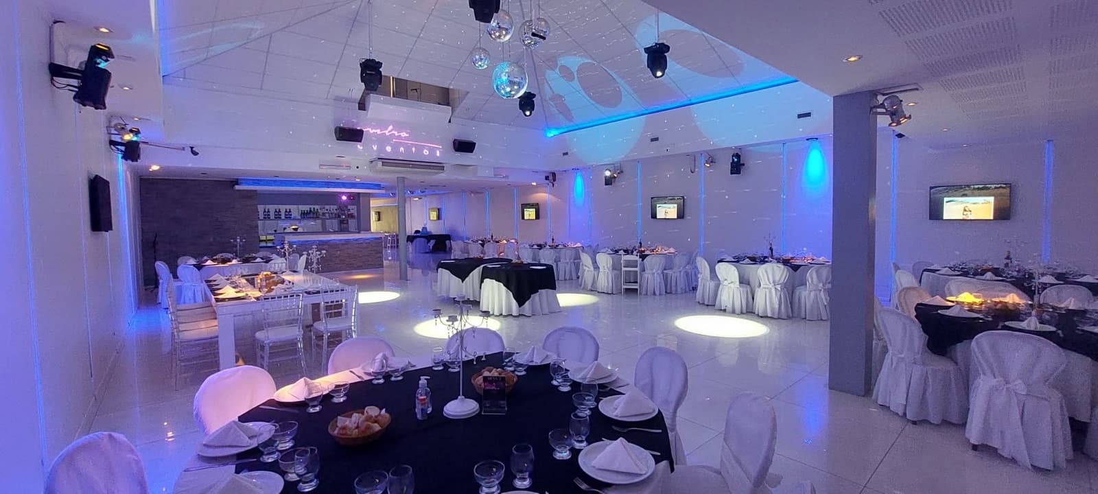 Panorámica del salón vacío con las mesas redondas puestas, las sillas con funda blanca y la luz azul en el techo.