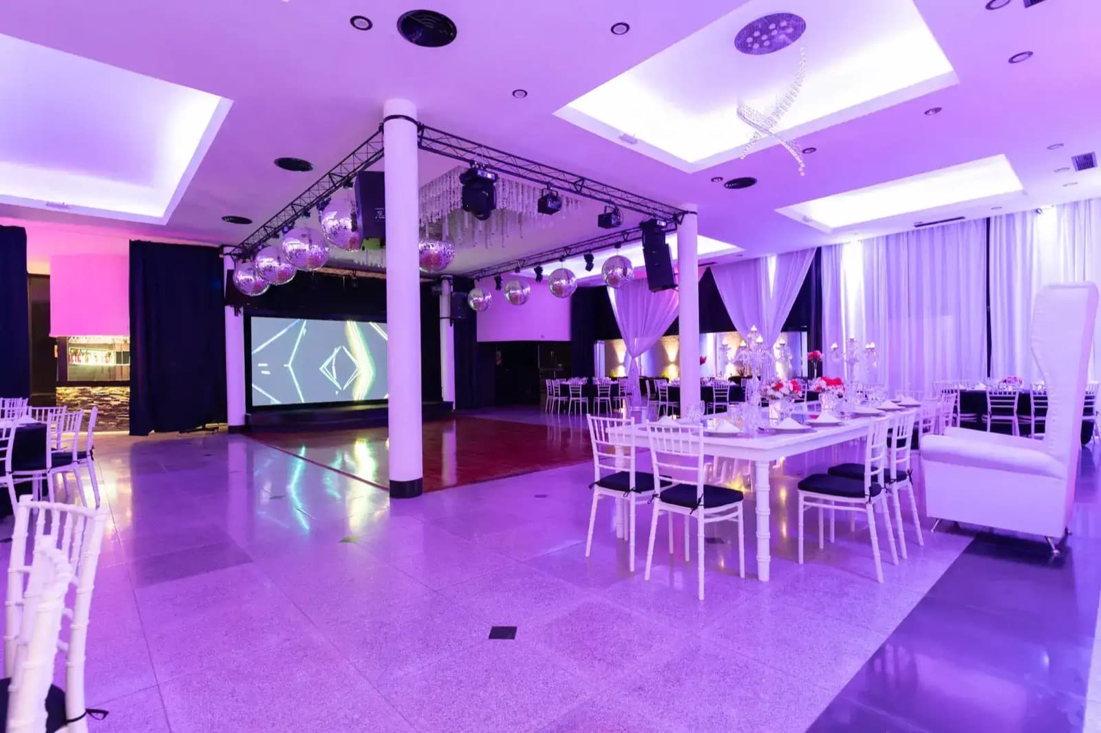 El salón Harmony puesto con mesas largas de mantel blanco y sillas Tiffany, esferas espejadas colgadas de una estructura de truss sobre la pista de parquet y una pantalla encendida al fondo, todo bañado de luz violeta.