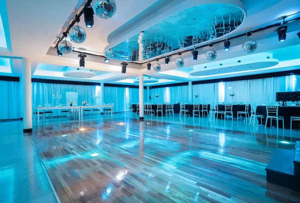 El salón Elegance vacío, con la pista de parquet lustrado en el centro, mesas de mantel negro y sillas plateadas contra cortinados blancos, y esferas espejadas bajo un cielorraso con espejos.