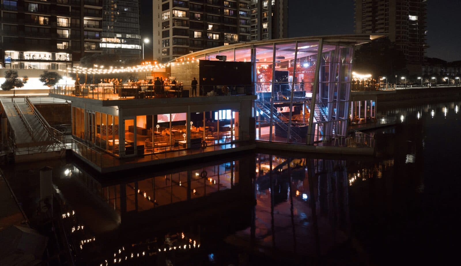 El salón flotante de noche desde la pasarela, con las terrazas iluminadas de cálido, los ventanales espejándose en el agua del dique y las torres de Puerto Madero detrás.