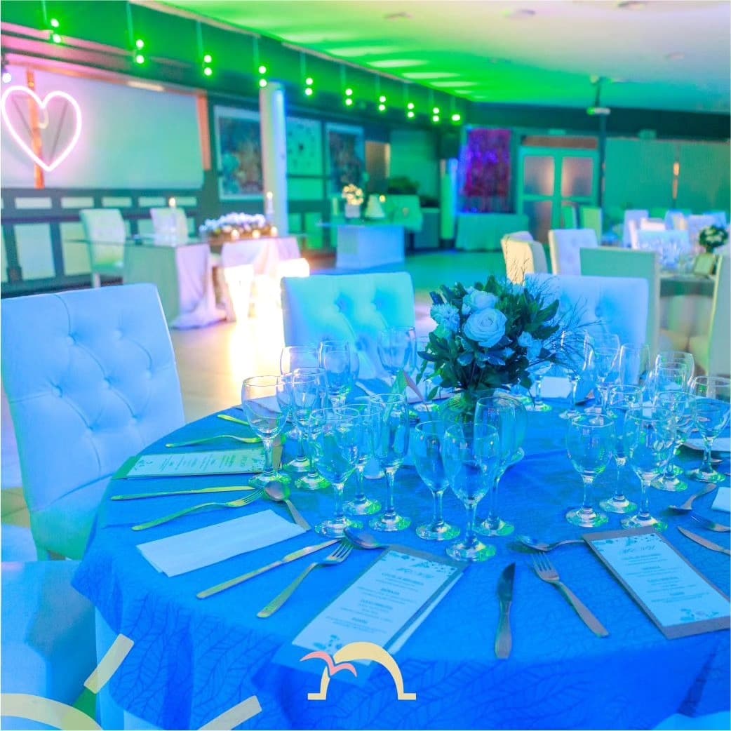 El salón puesto para una fiesta, con una mesa redonda de mantel celeste en primer plano, copas y un centro de rosas blancas, y el resto del salón iluminado de verde y azul con un corazón de neón en la pared.