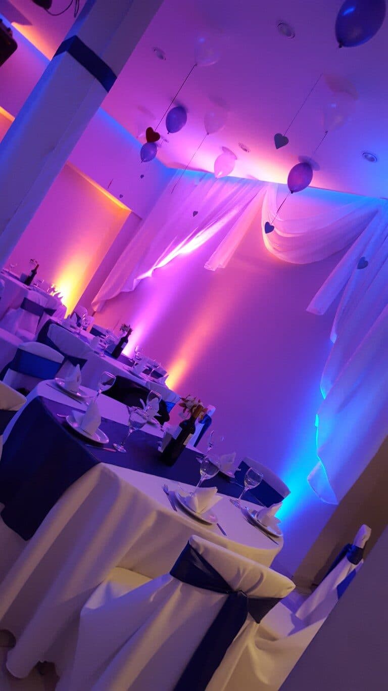 Las mesas vestidas de blanco con moños azules y copas servidas, bajo cortinados y globos, con las paredes bañadas de luz rosa y celeste.