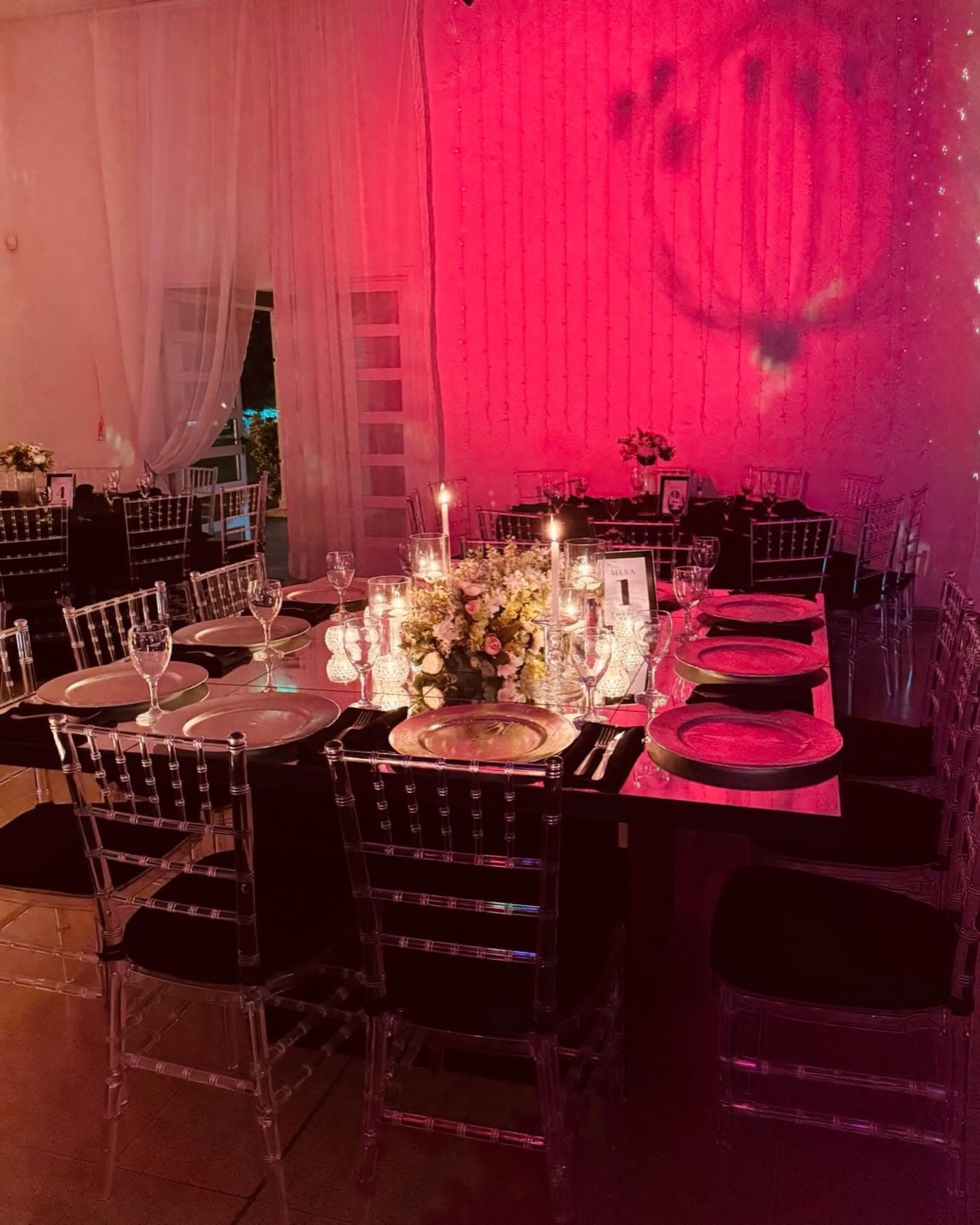 Una mesa larga con platos de presentación, velas y un centro bajo de flores, entre sillas transparentes, con la pared del fondo iluminada de fucsia y cortinados claros a un costado.