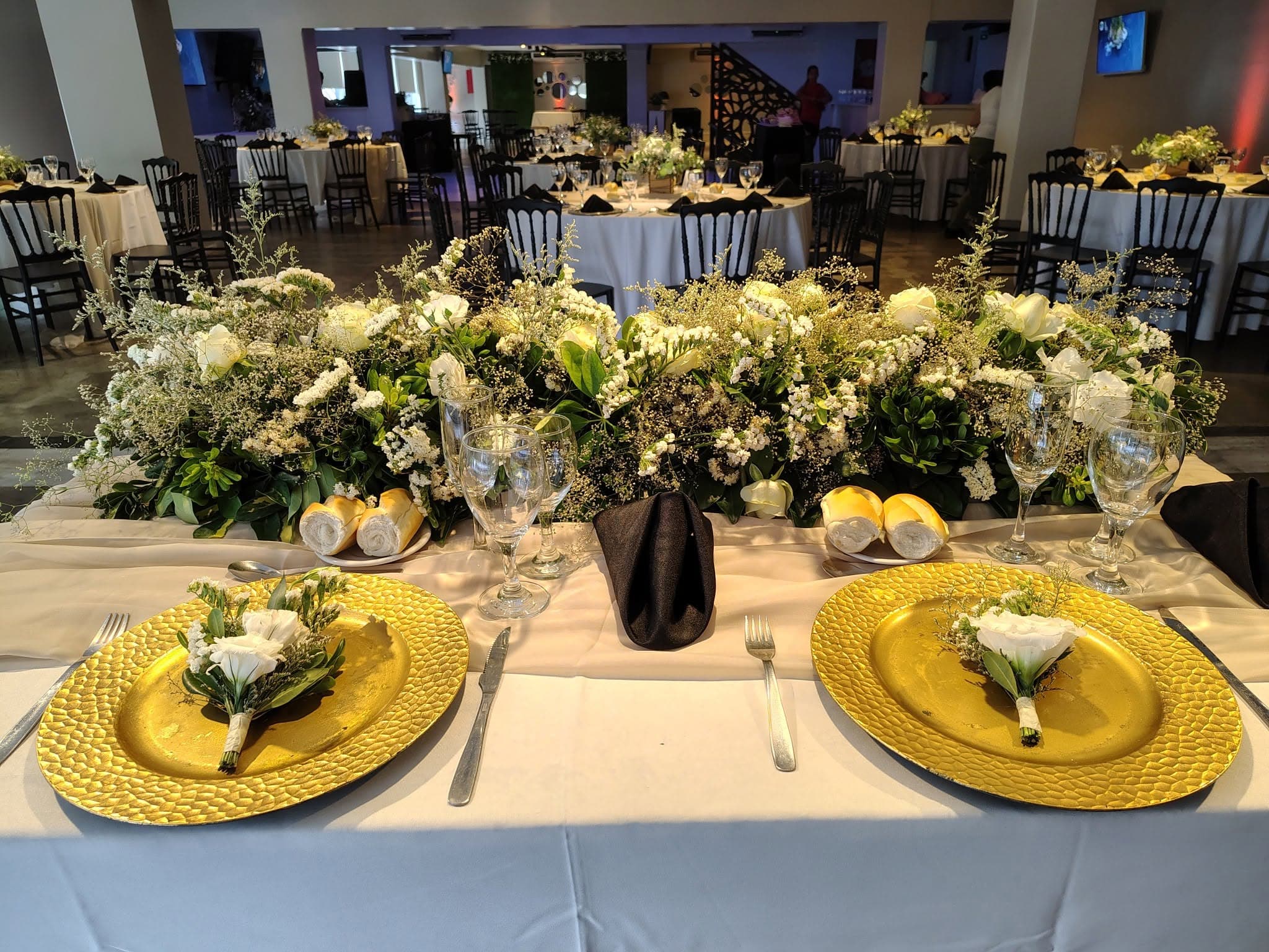 La mesa principal montada con platos dorados, copas y un centro largo de flores blancas y follaje, y detrás el salón con las mesas redondas vestidas de blanco y sillas negras.