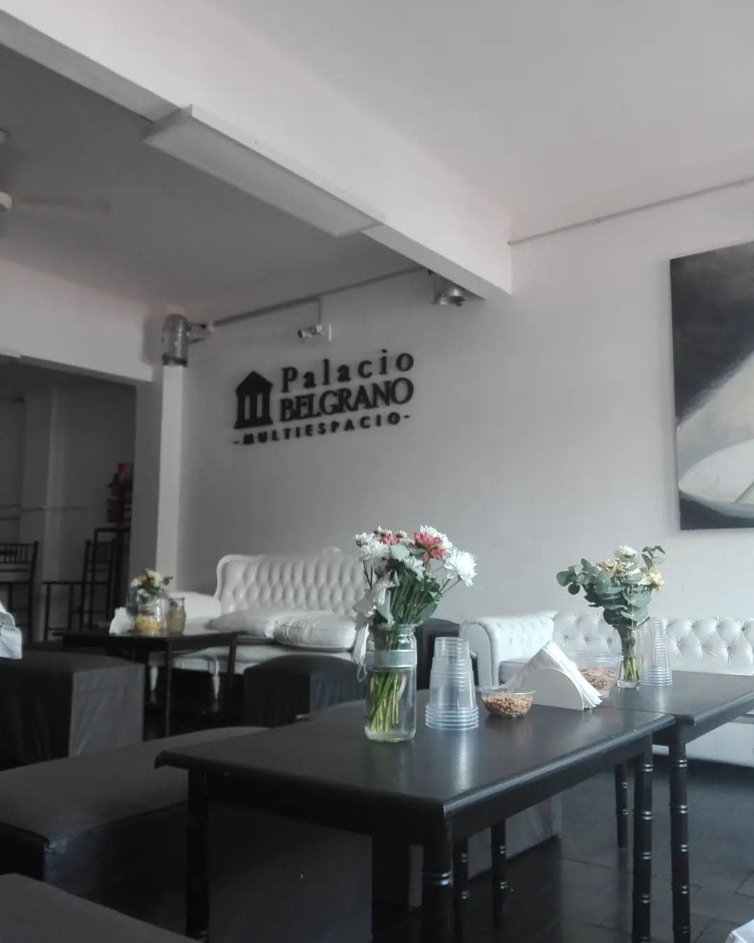 El interior del salón con el logo del lugar en relieve sobre la pared blanca, sillones capitoneados blancos contra el fondo y mesas negras con frascos de flores adelante.