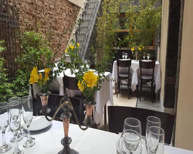 El patio con mesas vestidas de blanco, candelabro de plata y ramos de flores amarillas, contra la pared de ladrillo y la escalera de hierro del fondo.