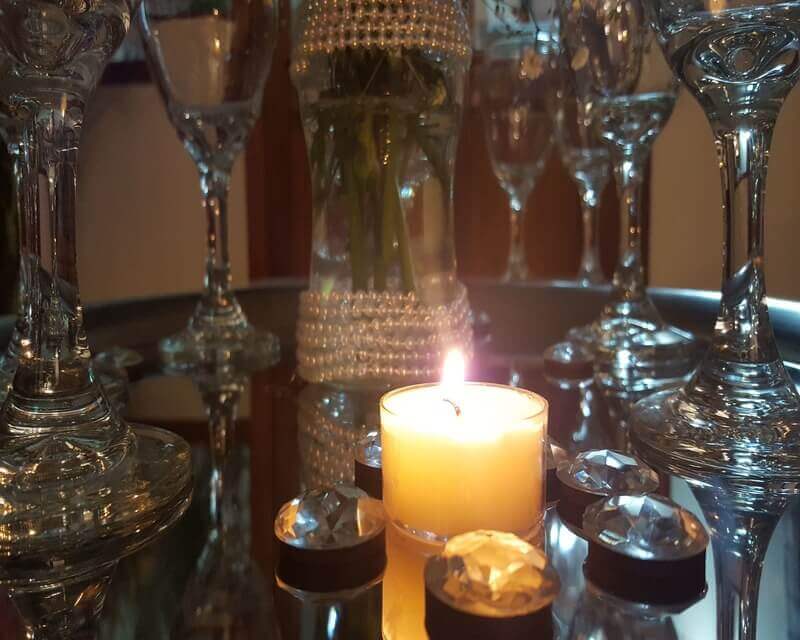 Detalle de una mesa espejada con copas altas, una vela encendida y piedras de cristal alrededor, con un florero forrado en perlas al fondo.
