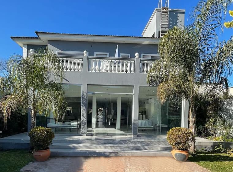 El frente del salón de día: una casa blanca de dos plantas con balaustrada, entrada vidriada, palmeras a los costados y un camino de baldosas hasta la puerta.