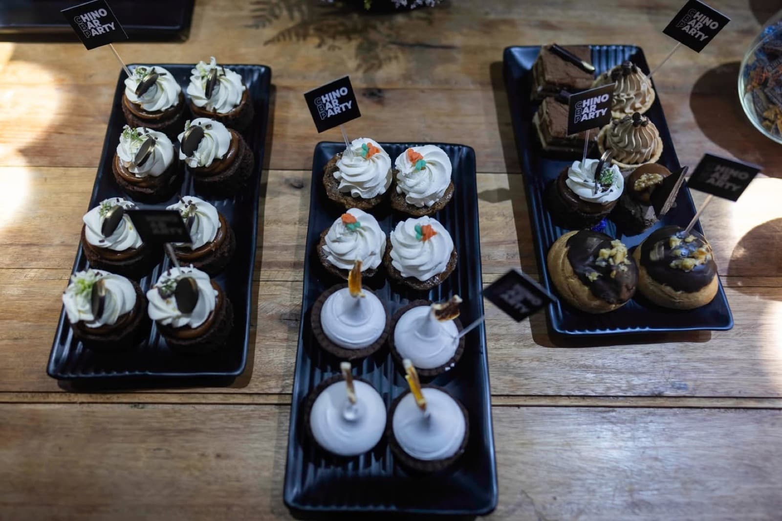 Bandejas negras con postres individuales sobre una mesa de madera, cada una con su cartelito.