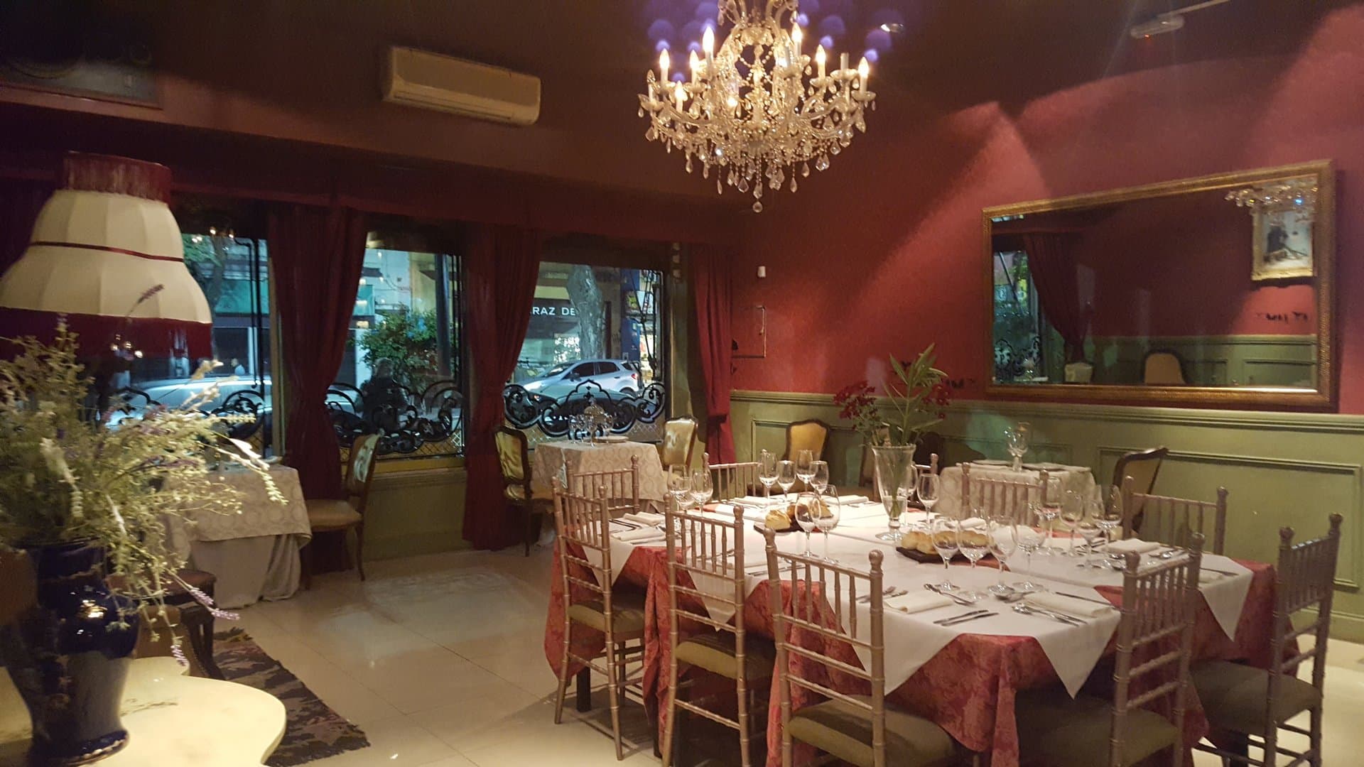 El salón vestido de noche, con araña de caireles, paredes bordó y verdes, espejo dorado y una mesa larga con mantel blanco sobre bordó y copas servidas.
