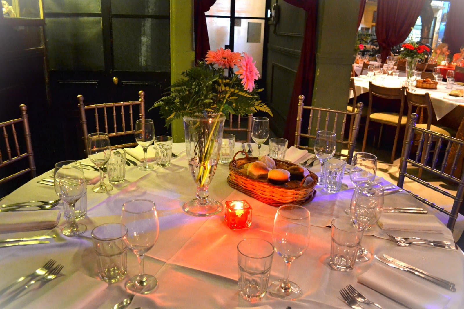 Una mesa redonda servida con copas, cubiertos y servilletas blancas, un ramo de gerberas rosadas en el centro, una vela encendida y la canasta de pan.