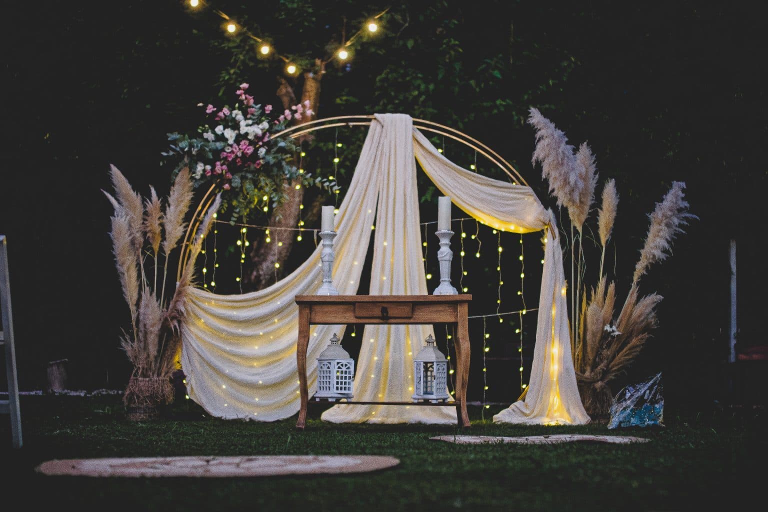 El arco de ceremonia de noche sobre el césped, con telas blancas, guirnaldas de luces, plumas de pampa a los costados y una mesa de madera con faroles en el centro.