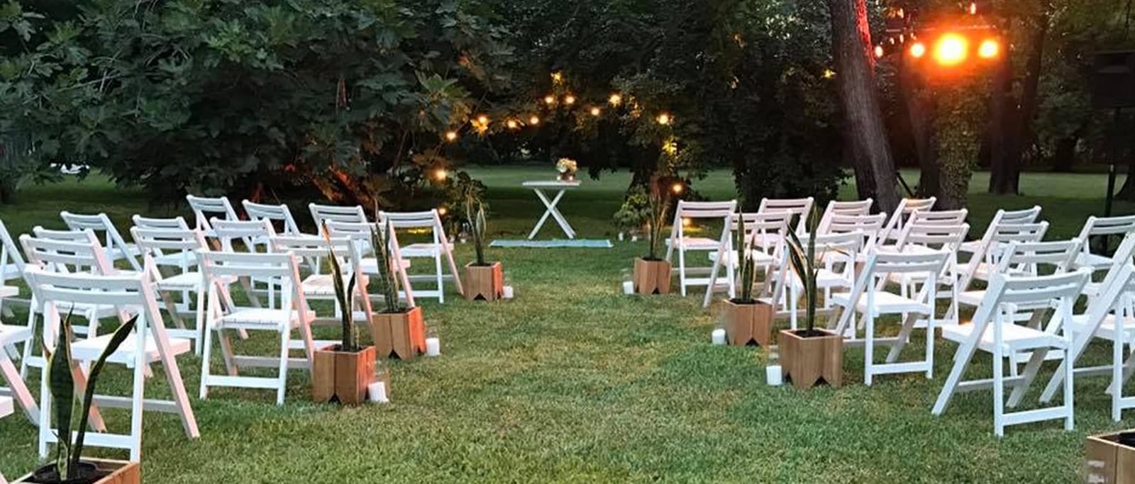 Las sillas blancas de la ceremonia alineadas sobre el césped de noche, con faroles en el piso y guirnaldas entre los árboles.