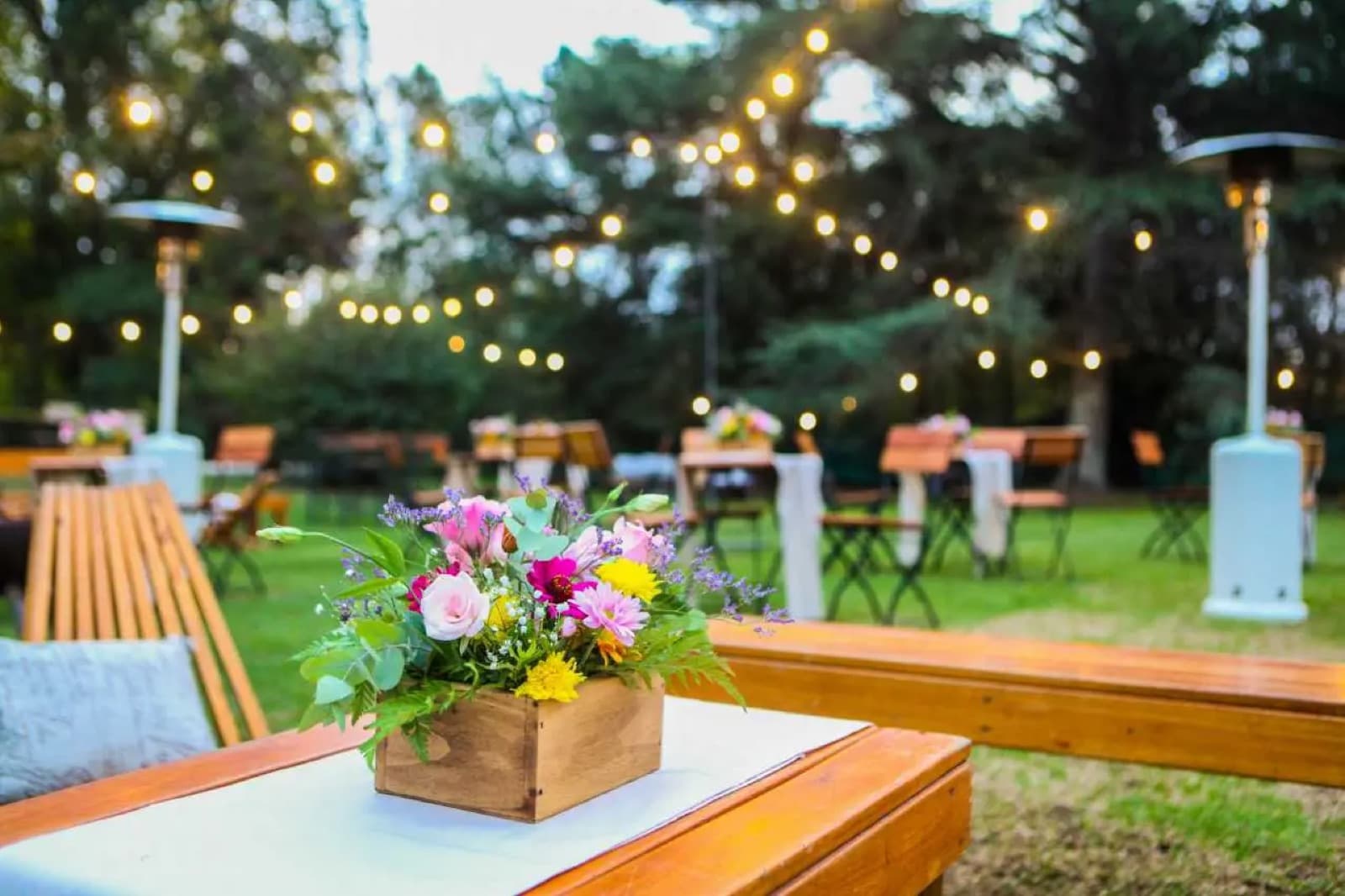 Un cajón de flores sobre un banco de madera en el parque, con las guirnaldas de luces y las estufas de exterior detrás.