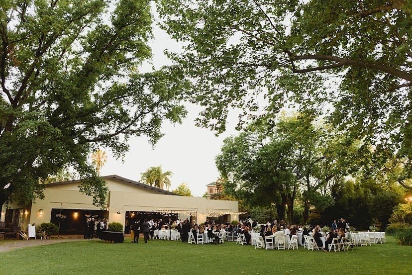 La fiesta montada en el parque al atardecer, con las mesas largas bajo los árboles y el salón de techo a dos aguas con guirnaldas al fondo.