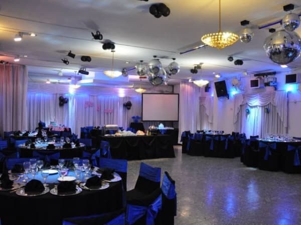 El salón armado con mesas redondas de mantel negro y sillas con moños azules, bolas de espejo y luces de colores colgando del techo, y una pantalla de proyección sobre el cortinado blanco del fondo.