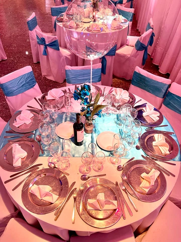 Una mesa redonda servida con mantel blanco, camino celeste, platos de vidrio y copas, con un globo transparente de centro de mesa y, atrás, más mesas con sillas vestidas de blanco y moños azules sobre el piso granítico.