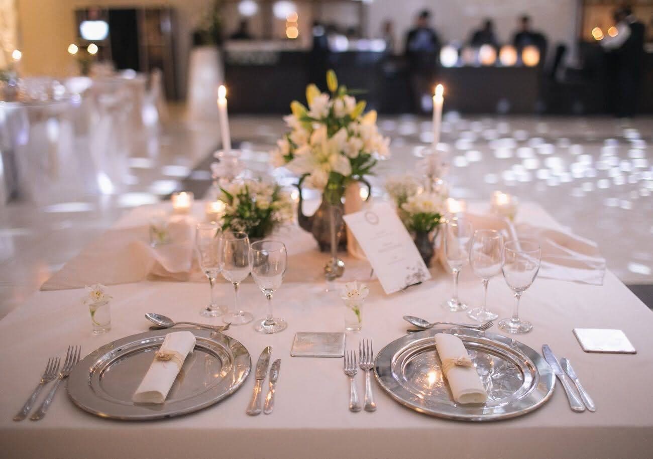 Una mesa puesta con mantel claro, platos con servilleta doblada, copas, velas encendidas y arreglos de flores blancas.