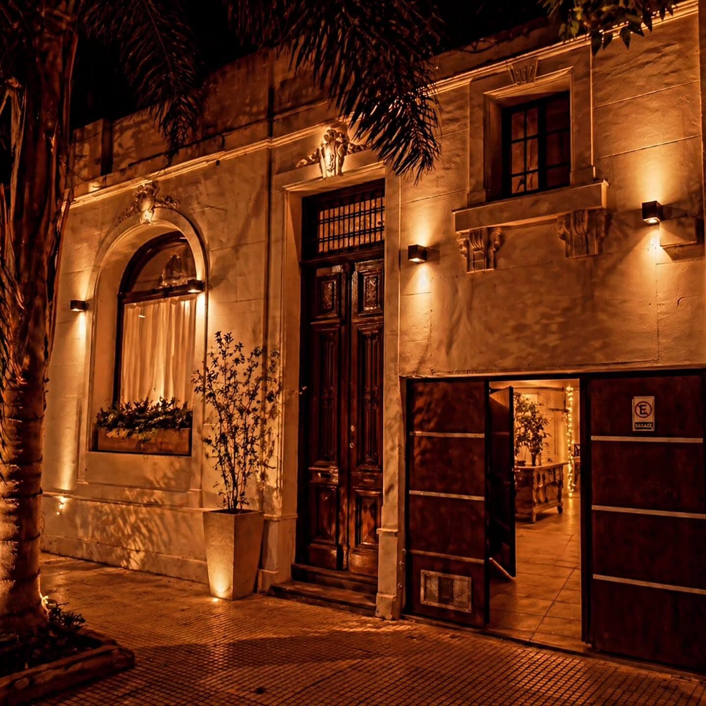 La fachada de la casa de noche, con la puerta de madera tallada iluminada, el portón del garage abierto dejando ver el interior con guirnaldas, y una palmera sobre la vereda de baldosas.