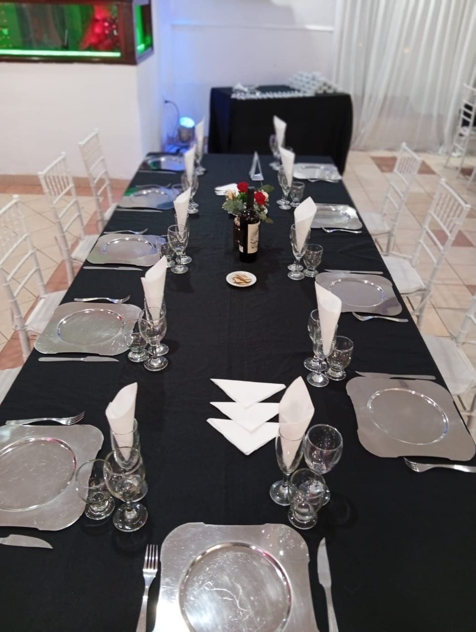 Una mesa larga vestida con mantel negro y platos de plata, servilletas paradas en cada lugar y una botella con flores rojas de centro, entre sillas blancas.