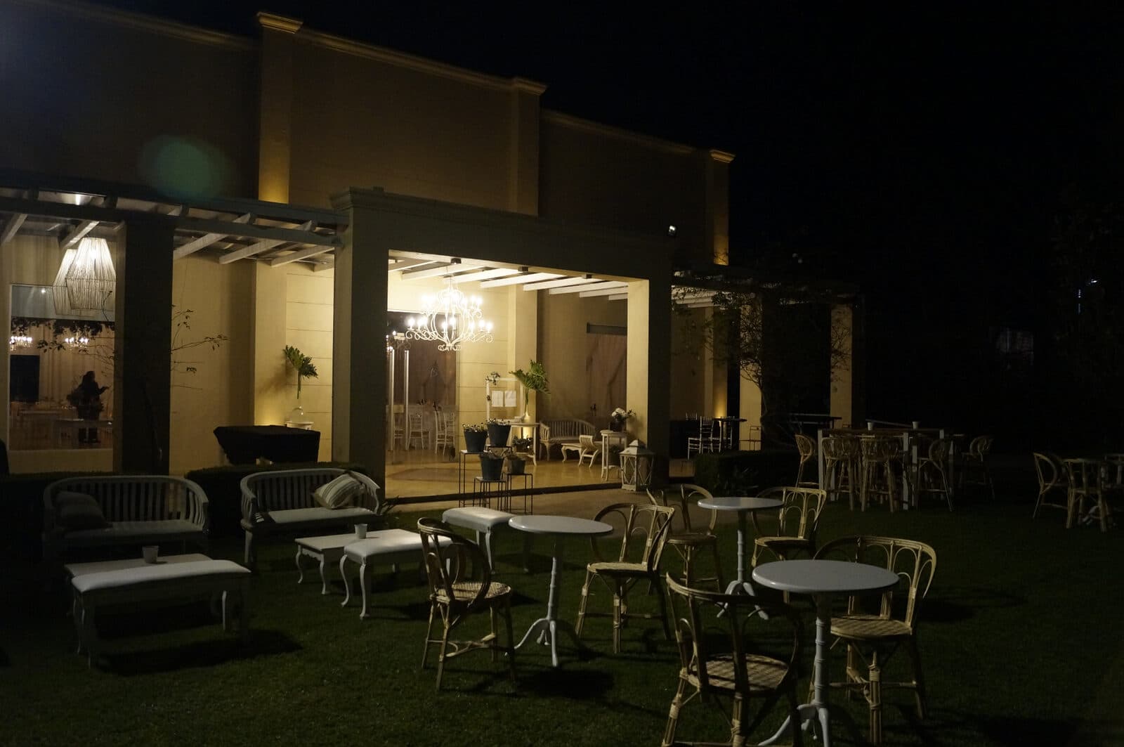 El frente del salón de noche, con las galerías iluminadas y mesas redondas con sillas armadas sobre el césped del parque.