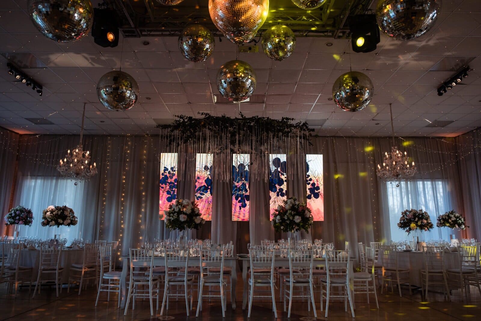 El salón armado para una fiesta, con bolas de espejo colgando del techo, cortinados claros, pantallas encendidas y mesas con sillas Chiavari blancas.
