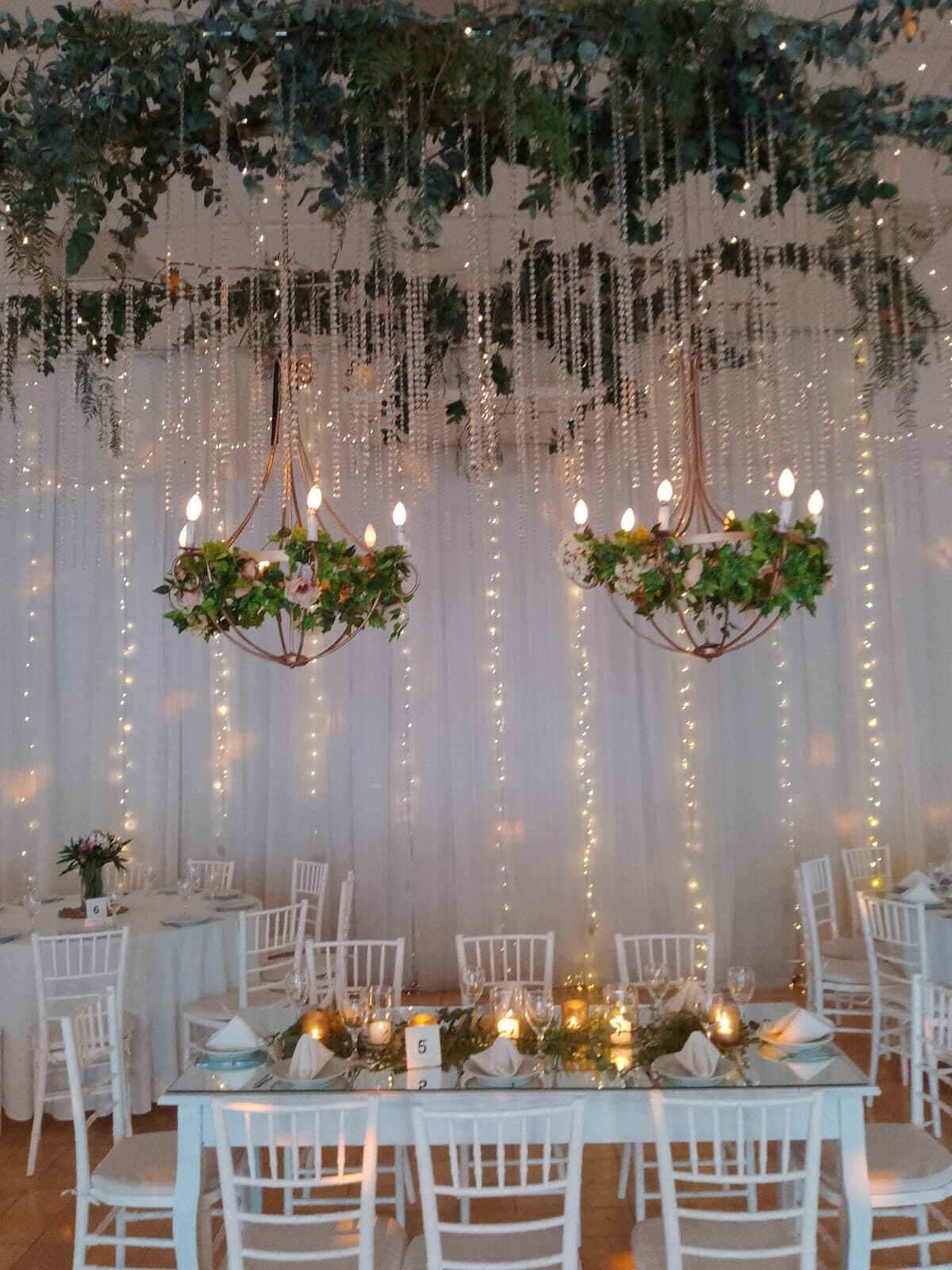 Una mesa larga bajo una estructura con enredaderas colgantes y arañas de luz, con sillas blancas y centros de plantas.