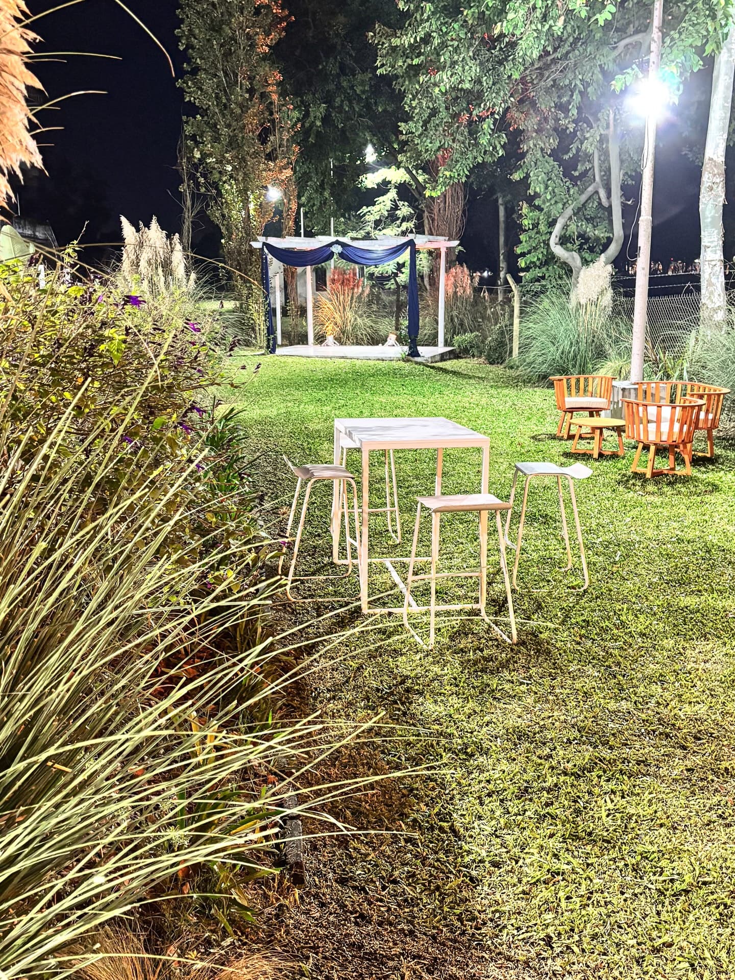 El parque de noche iluminado, con mesas altas y banquetas sobre el césped, sillones de mimbre a un costado y al fondo el altar de la ceremonia con cortinados azules.