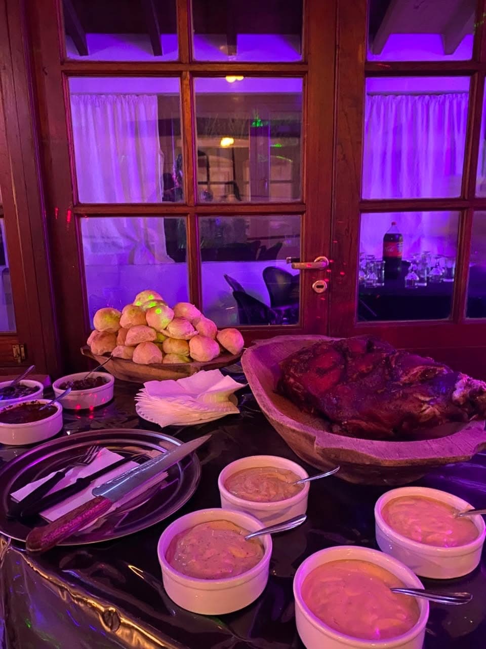 La mesa del bufet servida contra los ventanales de madera del salón, con una fuente de carne asada, una bandeja de pancitos y compoteras de salsas.