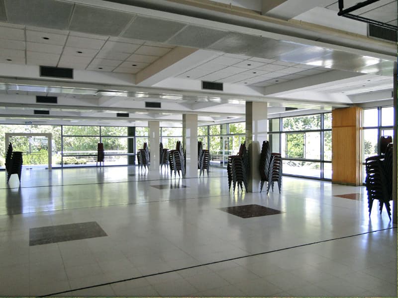 El salón de día y sin armar, con las sillas apiladas contra el vidriado que da al verde, las columnas blancas en el medio y el piso de cerámica reflejando la luz natural.