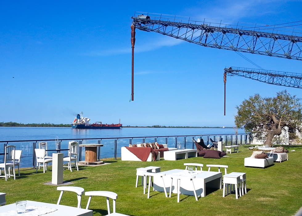 La terraza del salón sobre el río Paraná, con mesas y livings blancos sobre el césped, las grúas del puerto arriba y un buque pasando.