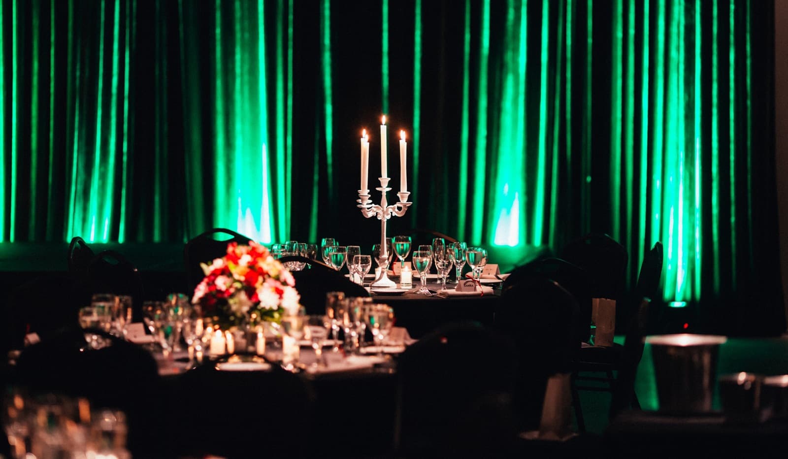 Una mesa larga puesta con candelabros y flores contra un cortinado iluminado de verde.