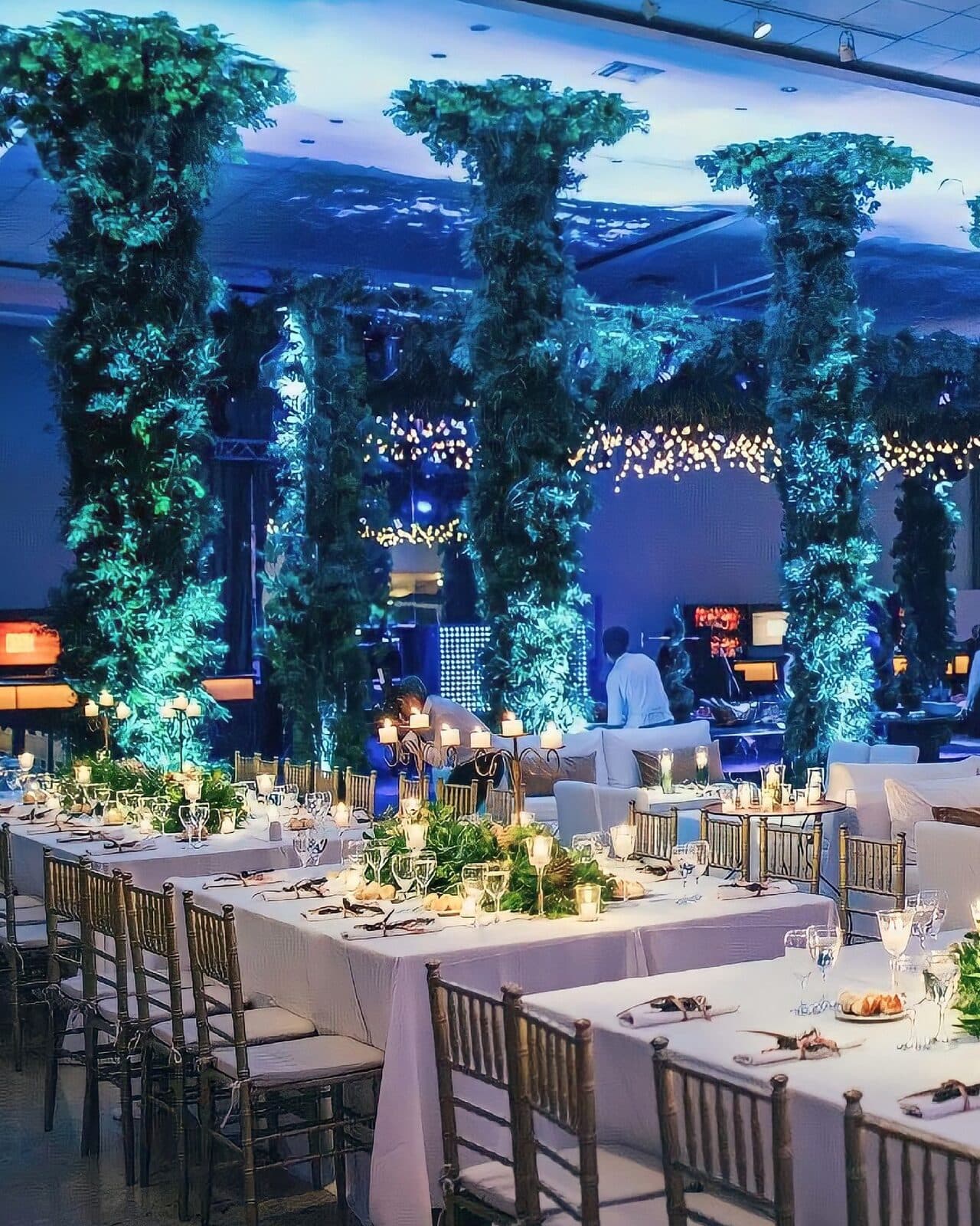 Mesas largas vestidas de blanco con candelabros y guías de follaje, sillas Chiavari doradas y columnas cubiertas de plantas iluminadas de verde bajo un cielorraso proyectado.