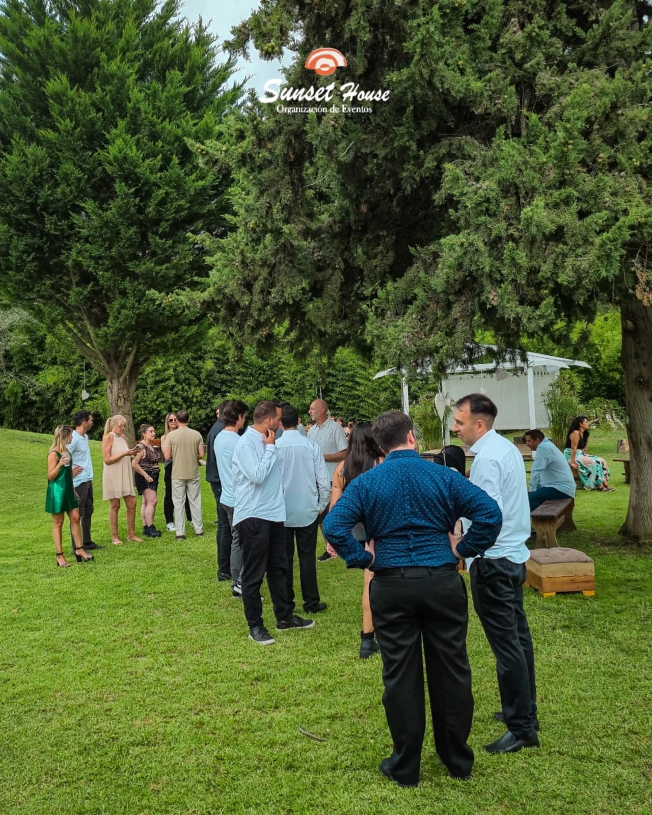 El parque de la quinta con cipreses altos, invitados de pie sobre el césped y la pérgola blanca a un costado.