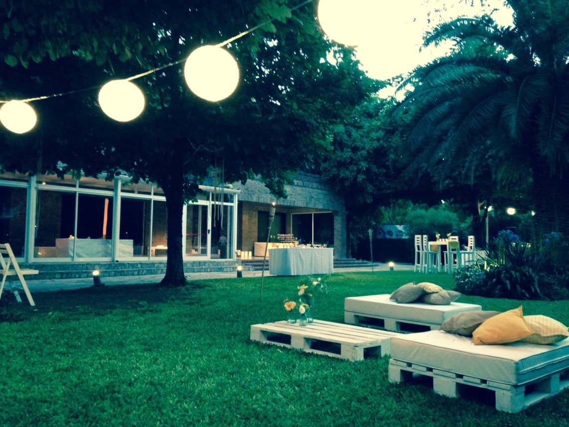 El parque al anochecer con farolas de papel colgadas entre los árboles, livings de pallet con almohadones y el salón vidriado iluminado al fondo.