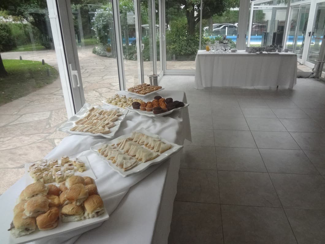 La mesa de recepción con bandejas de sándwiches y facturas, contra los ventanales que dan al parque y la pileta.