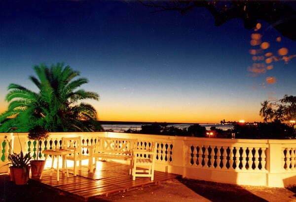 La terraza al atardecer, con la baranda de balaustres, sillas y macetas, y el río y el cielo naranja de fondo.