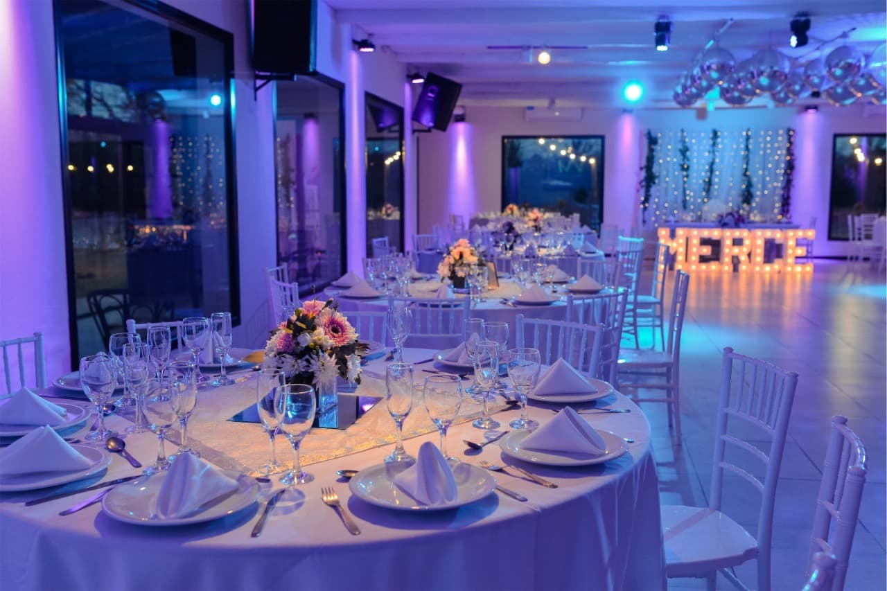 El salón puesto para una fiesta, con mesas redondas de mantel blanco, sillas Tiffany y la pista al fondo bajo las bolas de espejo.