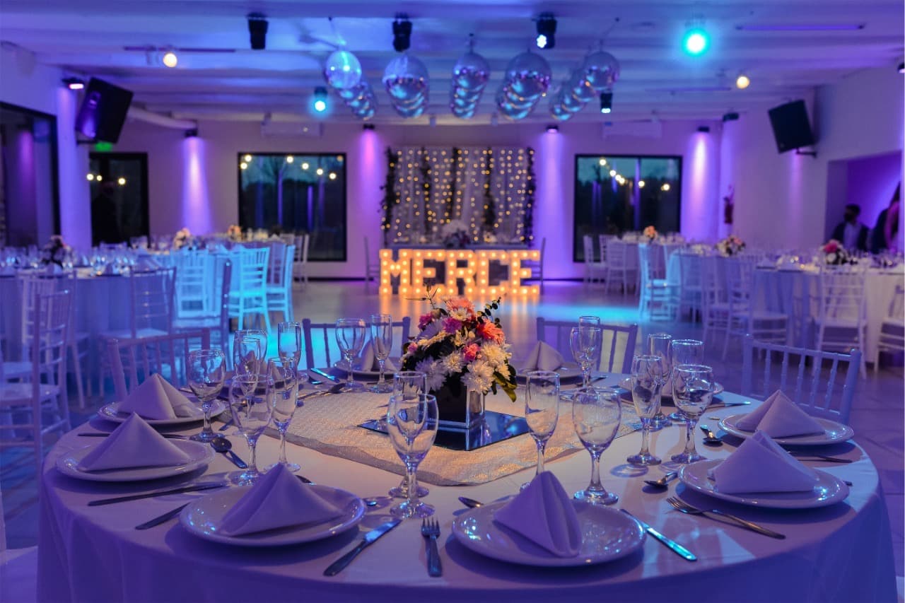 Una mesa tendida en primer plano con copas y servilletas, y detrás el salón entero iluminado en violeta con las letras luminosas del nombre.