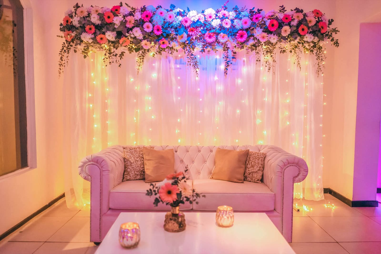 El living de la entrada, con un sillón capitoné, mesa baja con velas y un arco de flores sobre una cortina de luces.