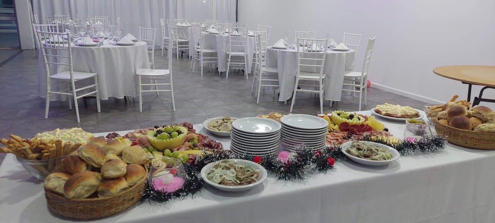 El salón puesto para una fiesta, con mesas redondas vestidas de blanco al fondo y una mesa larga de fiambres y panes en primer plano.